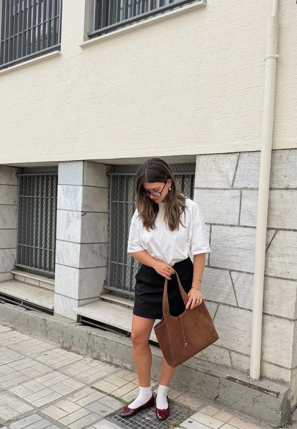 Kvinde står på en brolagt gade iført hvid skjorte, sort nederdel, hvide sokker, bourgogne sko og holder en brun håndtaske nær en bygningsvæg.
