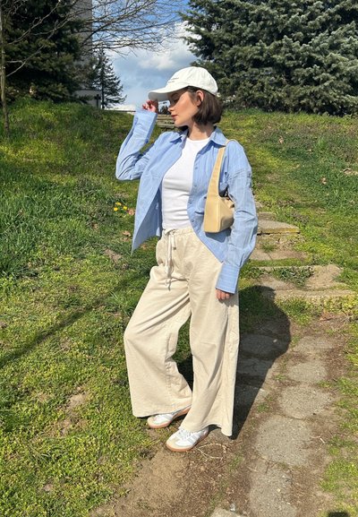 Camisa de botón azul claro sobre una camiseta blanca, pantalones de pierna ancha beige y zapatillas blancas. Una gorra crema y un bolso beige completan el look.