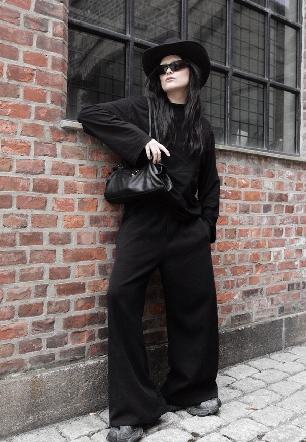 Person trägt weite schwarze Hose, schwarzes Oberteil, schwarzen Hut, Sonnenbrille und hält eine schwarze Handtasche, lehnt an einer Backsteinmauer mit Fenstern.