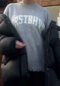 Personne portant un pull en maille gris avec le mot "FIRSTBHVF" et une doudoune noire, debout devant un mur en briques.