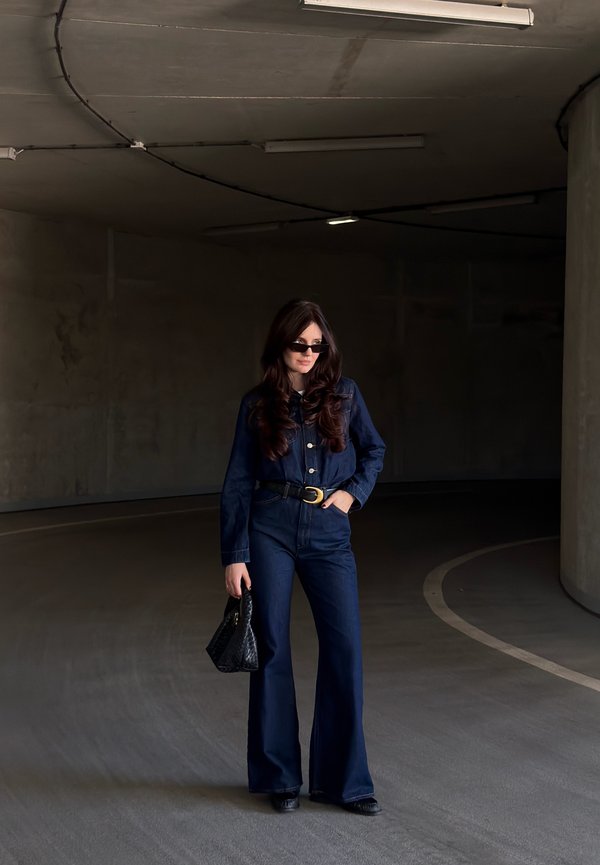 Mujer con mono vaquero azul oscuro y gafas de sol, de pie en un párking curvo de hormigón con poca luz, sosteniendo un bolso negro.