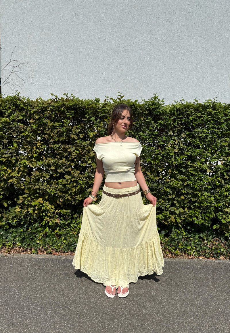 Femme en haut jaune clair décolleté aux épaules et jupe longue se tient sur le trottoir devant une haie verte, tenant les bords de la jupe avec les deux mains.