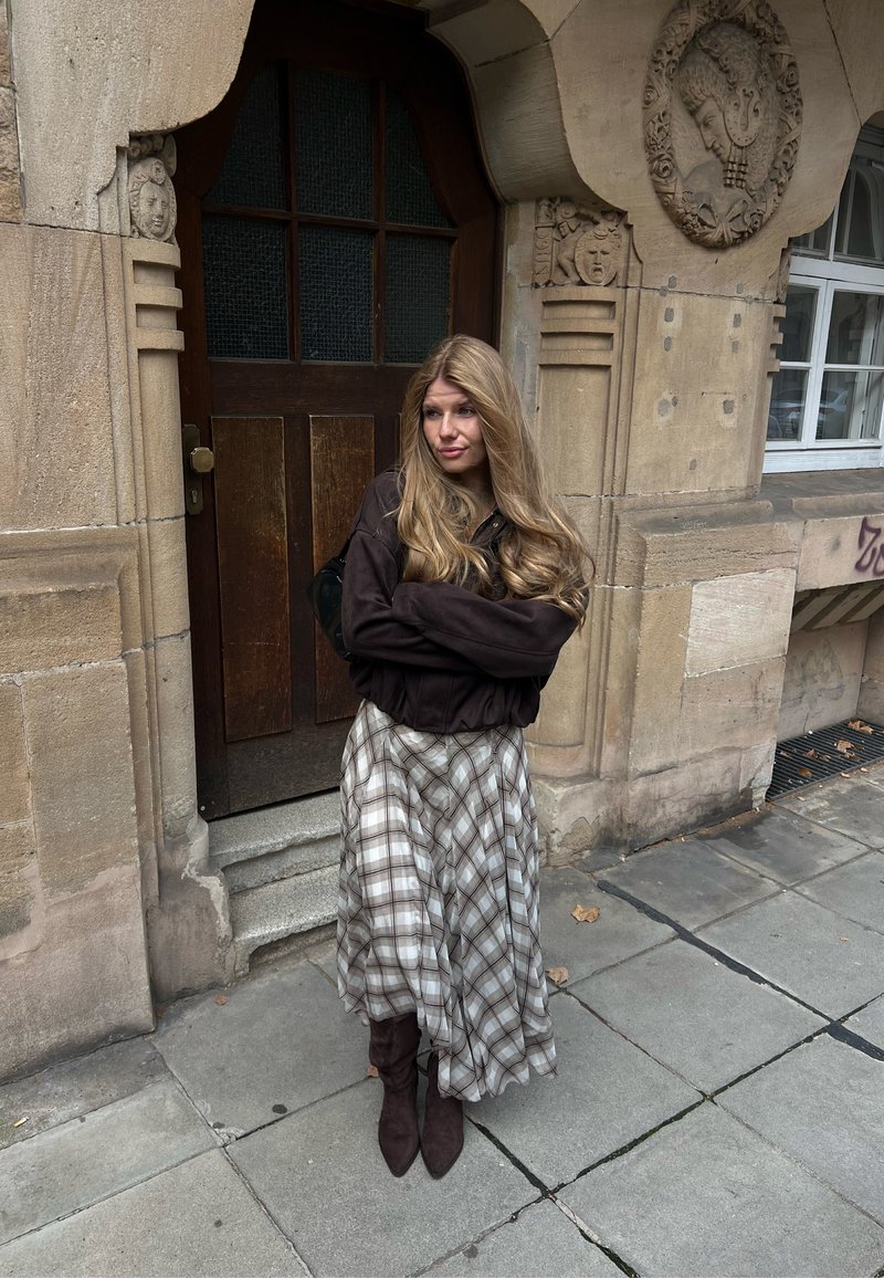 Une femme portant une veste sombre et une jupe à carreaux se tient les bras croisés devant une entrée en pierre ornée, dans une rue de la ville.