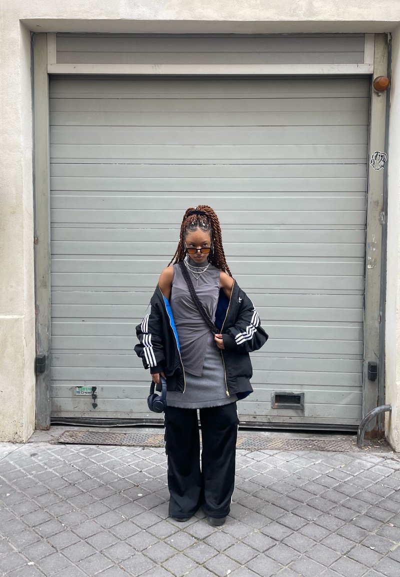 Μαύρο bomber μπουφάν με μπλε λεπτομέρειες, γκρι αμάνικο φόρεμα, φαρδιά μαύρα παντελόνια, καστανά πλεγμένα μαλλιά, μαύρα γυαλιά και ακουστικά.