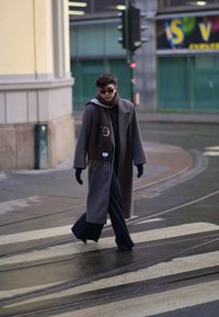 Casaco cinzentão sobre roupa negra, grandes óculos de sol escuros, luvas escuras e um cachecol castanho com franjas, a caminhar num passadeira molhada.