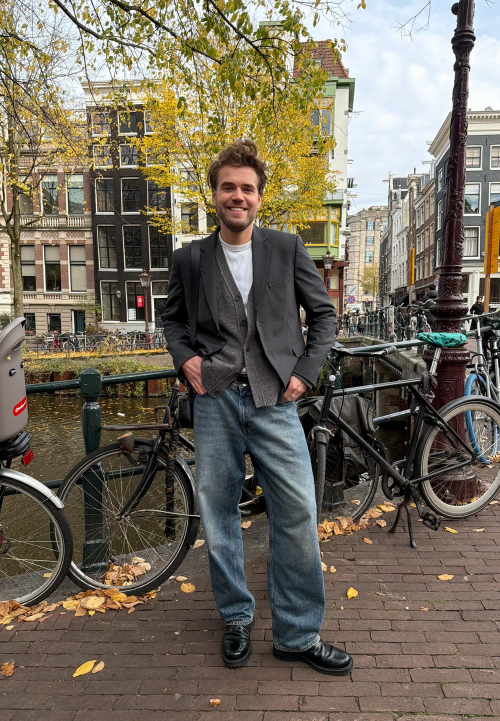 Homem com blazer preto sobre um cardigã cinza, t-shirt branca e jeans azul claro, em pé perto de bicicletas junto a um canal com folhas de outono.