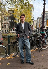 Homem com blazer preto sobre um cardigã cinza, t-shirt branca e jeans azul claro, em pé perto de bicicletas junto a um canal com folhas de outono.