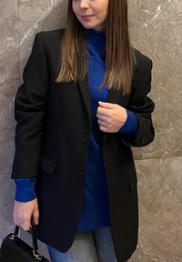 Čierne oversize sako s dvoma gombíkmi, doplnené modrým rolákom a sivými džínsami. Doplnky zahŕňajú malú čiernu kabelku.