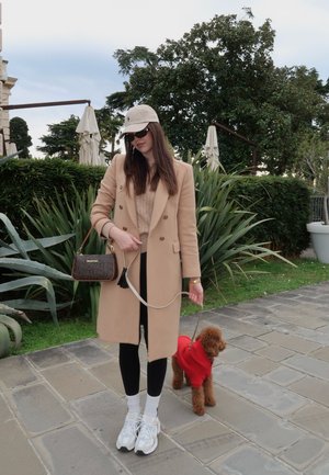 Mujer con abrigo beige, sombrero y gafas de sol sosteniendo la correa de un perro pequeño marrón que lleva un suéter rojo en un camino pavimentado.