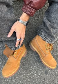 Stivali in suede marrone chiaro con punta rinforzata, lacci gialli e suole in gomma. Indossati con jeans grigi e una giacca di pelle bordeaux.