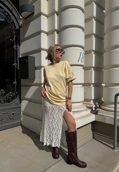 Mujer con cabello rubio y gafas de sol, vistiendo una blusa amarilla, una falda de encaje blanco con una abertura en el muslo y botas marrones hasta la rodilla, de pie junto a un edificio de piedra clara.