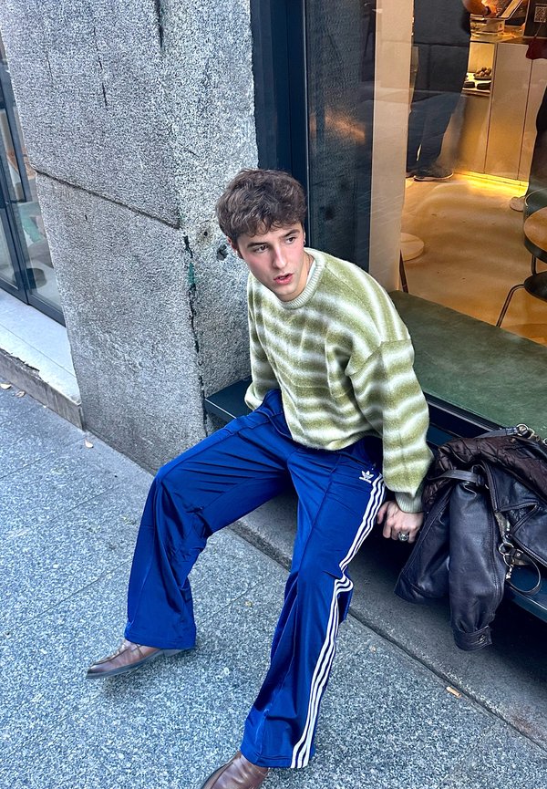Un maglione a righe verdi e bianche, pantaloni da jogging blu con righe bianche, scarpe marroni e una borsa in pelle marrone, seduto contro un muro di pietra.