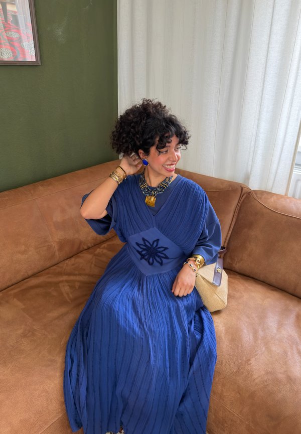 Femme aux cheveux bouclés portant une robe bleue et des bijoux remarquables, assise sur un canapé marron, souriante et se touchant les cheveux.