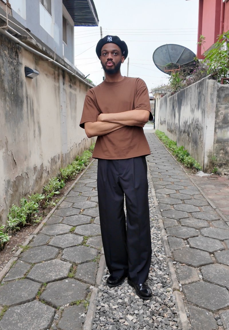 Chemise marron à manches courtes, pantalon noir ample et chaussures noires. En arrière-plan, un chemin étroit bordé de murs et de verdure.