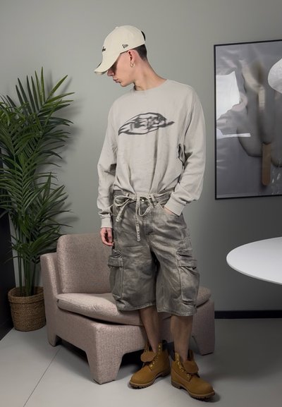 Camisa de manga larga beige con diseño gráfico, pantalones cortos cargo grises y botas marrones. De fondo hay una planta verde y una pared de color gris claro.