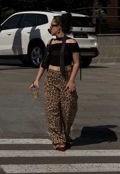 Top negro sin hombros con un cuello atado, pantalones sueltos con estampado de leopardo y zapatos rojos. Sosteniendo una bebida helada, caminando por un paso de peatones.