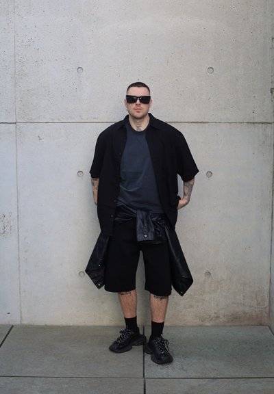 Camisa negra de botones sobre una camiseta oscura, pantalones cortos negros y zapatillas negras. Presenta tatuajes en los brazos y las piernas, gafas de sol y superficies texturizadas.
