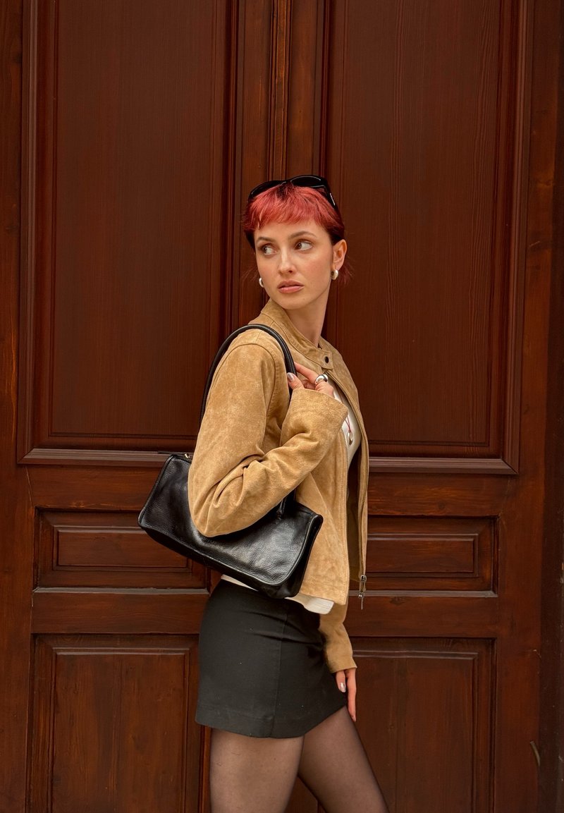 Joven con el pelo corto y rojo, lleva una chaqueta marrón claro, una falda negra y un bolso negro, de pie junto a una puerta de madera.