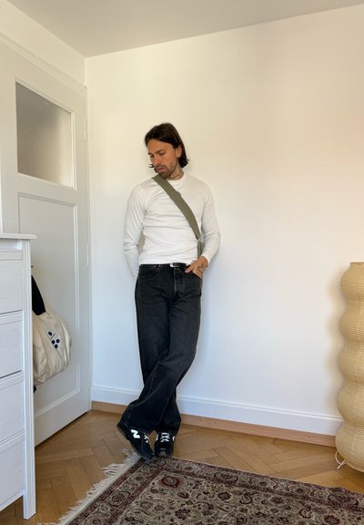 Hombre con camisa blanca de manga larga y vaqueros negros con bolso bandolera se apoya en una pared blanca en una habitación con suelo de madera y alfombra estampada.