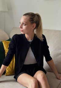 Čierny cardigan s gombíkmi a dlhými rukávmi, ktorý má na sebe šedý top, skombinovaný s čiernymi plisovanými šortkami. Sedí na svetlej pohovke s vankúšmi.