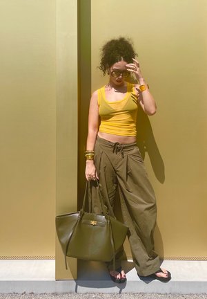 Top corto sin mangas amarillo, pantalones de pierna ancha verde oliva, un gran bolso de tote de cuero verde oscuro y gafas de sol contra un fondo dorado.