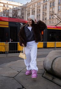 Ružičaste tenisice, bijele trenirke, sivi duks s patentnim zatvaračem i smeđi krzneni kaput. Usklađena žuta torbica. Urbani ambijent s tramvajem.