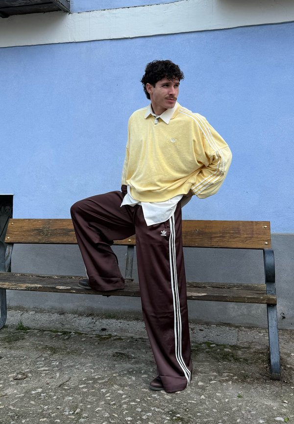 Man met krullend haar die een gele trui en bruine Adidas trainingsbroek draagt, staat met één voet op een houten bankje tegen een blauwe muur.