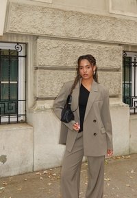 Blazer gris de gran tamaño y pantalones anchos a juego, combinados con una camiseta negra y un bolso negro texturizado, sobre un fondo de pared texturizada.