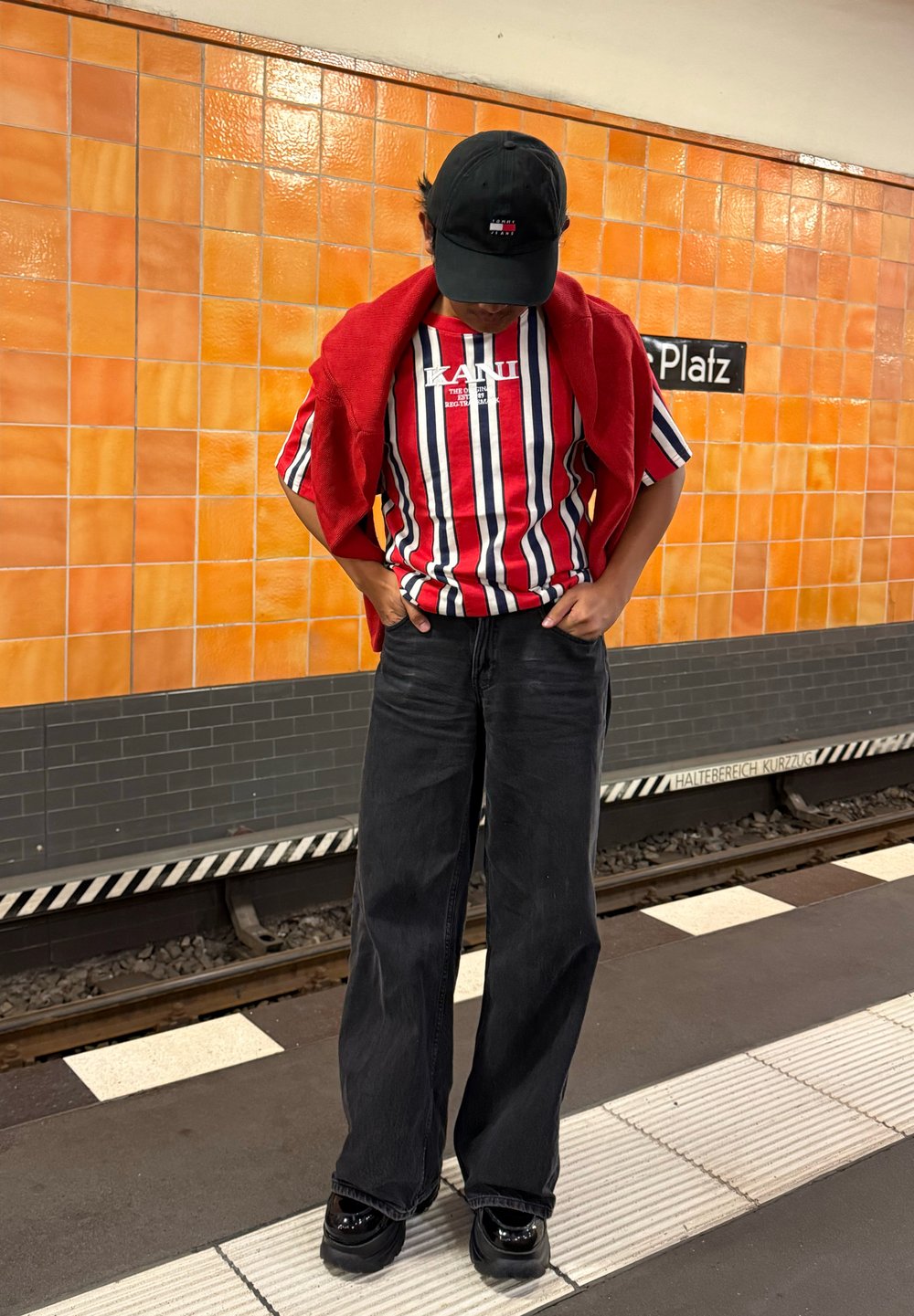 Person som har på seg en stripete rød, hvit og blå skjorte med en rød genser over skuldrene og svart caps, som står på en t-banestasjon.
