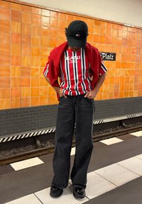 Person som har på seg en stripete rød, hvit og blå skjorte med en rød genser over skuldrene og svart caps, som står på en t-banestasjon.
