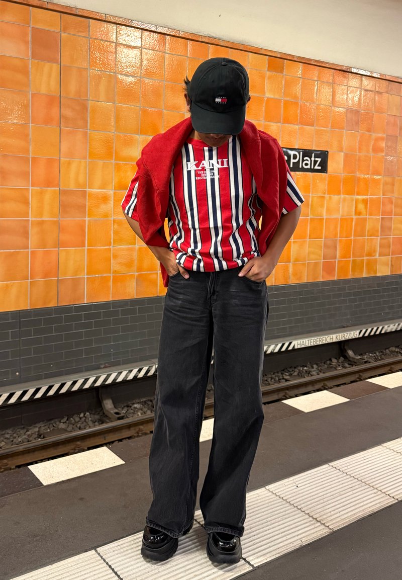 Persona che indossa una maglietta a righe rossa, bianca e blu con un maglione rosso sulle spalle e un berretto nero, in piedi su una banchina della metropolitana.
