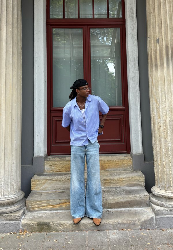 Camisa de manga corta azul clara sobre una camiseta blanca, combinada con jeans de pierna ancha descoloridos y tacones marrones de punta, con una puerta roja de fondo.