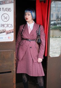 Une femme se tient dans une cabine photo, portant un costume à carreaux rose avec une taille cintrée, une chemise blanche et une cravate rose. Elle a les cheveux colorés et un sac à main noir.