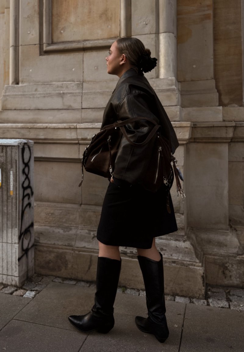 Woman walking on city pavement wearing a black leather jacket, black skirt, knee-high black boots, and carrying a large dark brown handbag.