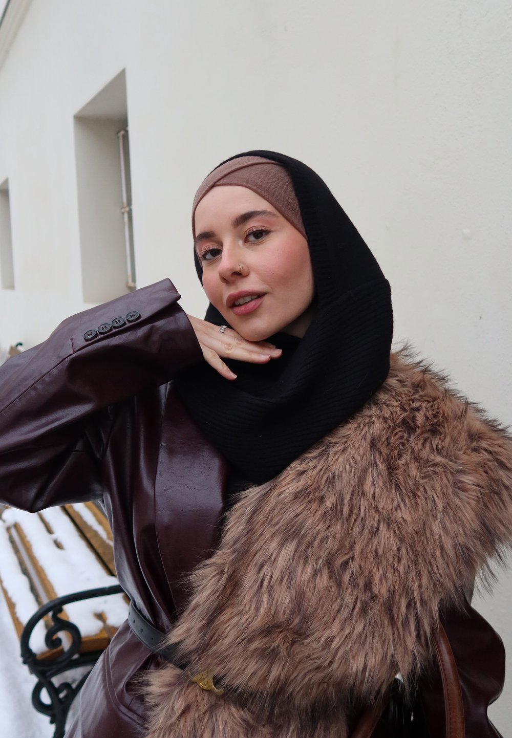 Femme portant un hijab avec un manteau en cuir et une écharpe en fourrure, posant avec la main près du visage devant un mur clair en extérieur.