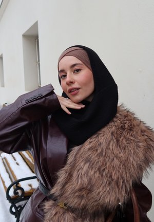 Mujer con hijab, abrigo de cuero y bufanda de piel posando con la mano cerca del rostro frente a una pared clara al aire libre.