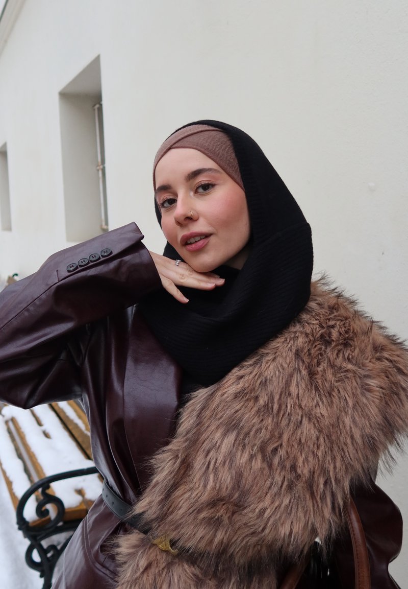 Kobieta w hidżabie, nosząca skórzany płaszcz i futrzany szalik, pozuje z ręką przy twarzy na tle jasnej ściany na zewnątrz.