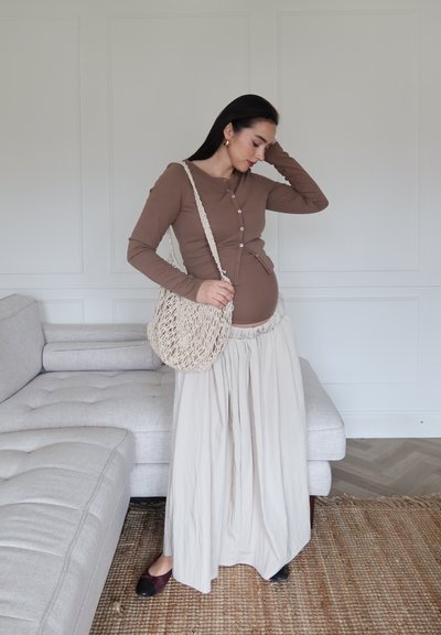 Top marrón de manga larga con botones, falda beige con cintura fruncida, bolso tejido beige, zapatos negros, fondo minimalista con sofá gris claro.