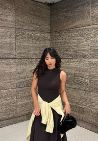 Brun tekstureret kjole med bådudskæring og ærmeløs design, kombineret med en gul cardigan med knude foran. Holder en lille sort pelsvaskepose.