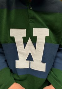 Grønn og blå genser med krage, som har en stor hvit "W"-logo foran. Stoffet har en glatt tekstur.