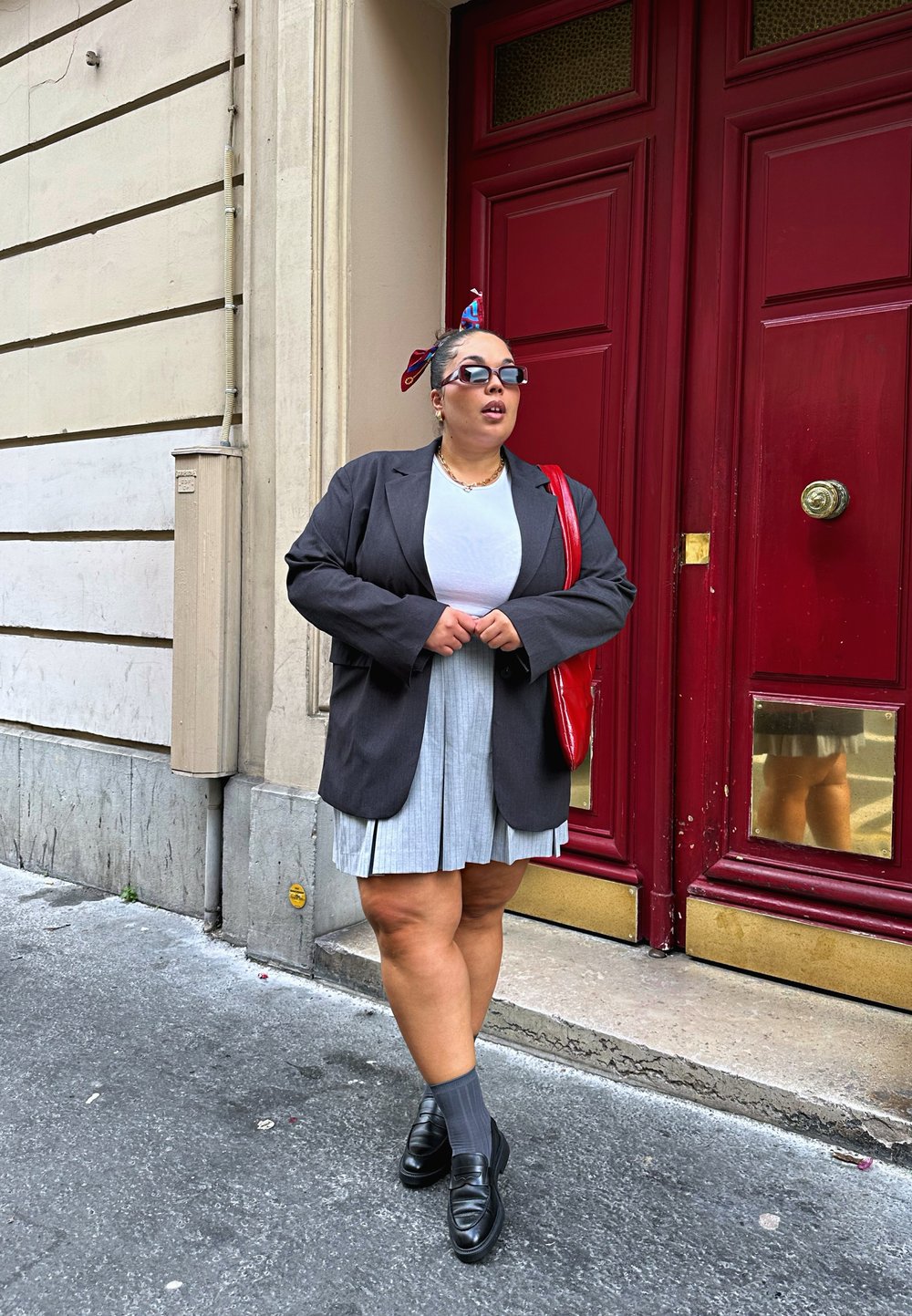 Γκρι πλισέ φούστα, λευκό μπλουζάκι, oversized μαύρο σακάκι και μαύρα παπούτσια. Κόκκινη τσάντα και κασκόλ με σχέδιο στα μαλλιά. Κόκκινη πόρτα στο φόντο.