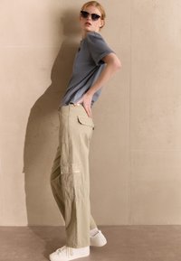 T-shirt grigia a maniche corte abbinata a pantaloni cargo beige con tasche laterali, indossata con sneakers bianche e occhiali da sole, su uno sfondo neutro.