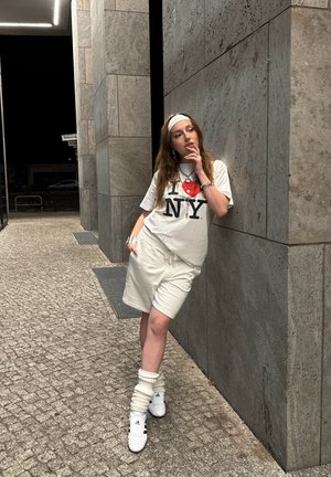 Modelo con una camiseta blanca "I love NY" y pantalones cortos beige, llevando zapatillas blancas con calentadores de piernas acanalados, frente a un fondo de pared de piedra.