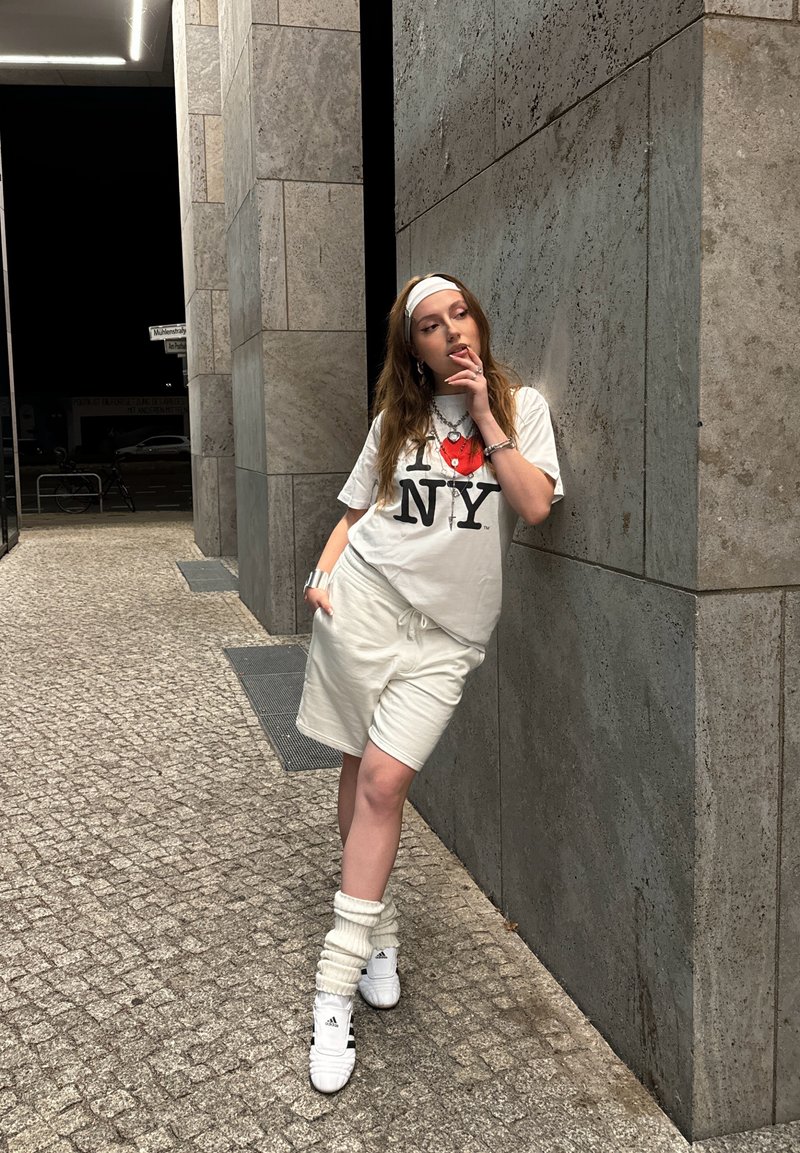 Λευκό T-shirt με γραφικό "I ❤️ NY", ανοιχτό μπεζ σορτσάκι, λευκά καλσόν ποδιών και λευκά αθλητικά παπούτσια μπροστά από τον τοίχο από πέτρες.