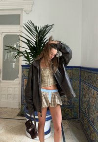 Καφέ δερμάτινο μπουφάν, καρό τοπ πουλάν, σορτς με μπλε λεπτομέρειες. Το φόντο περιλαμβάνει διακοσμητικά πλακάκια και πράσινο τοπίο. Εσωτερικός χώρος.