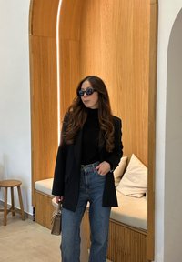 Μαύρο σακάκι πάνω από μαύρο λαιμόκοψη, συνδυασμένο με μπλε τζιν ψηλής μέσης. Τα αξεσουάρ περιλαμβάνουν σκούρα γυαλιά ηλίου και μια μικρή τσάντα. Φόντο από ξύλινα πάνελ.