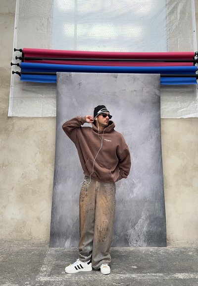 Hombre con sudadera con capucha marrón, pantalones anchos salpicados de pintura, gorro negro y zapatillas, de pie con una mano detrás del codo sobre un fondo texturizado con rollos de papeles de colores.