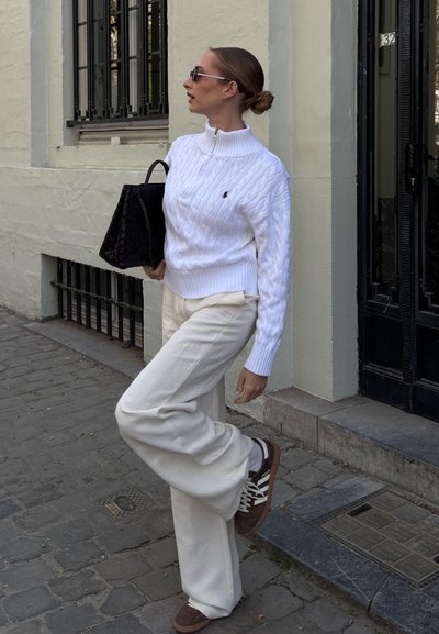 Suéter blanco de punto trenzado, pantalones de pierna ancha color blanco roto, zapatillas marrones, bolso tote negro, gafas de sol elegantes; de pie en una calle de adoquines.