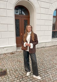 Blazer marron sur un col roulé blanc, pantalon large en cuir noir et baskets blanches. Sac bandoulière en daim marron. Sol pavé.