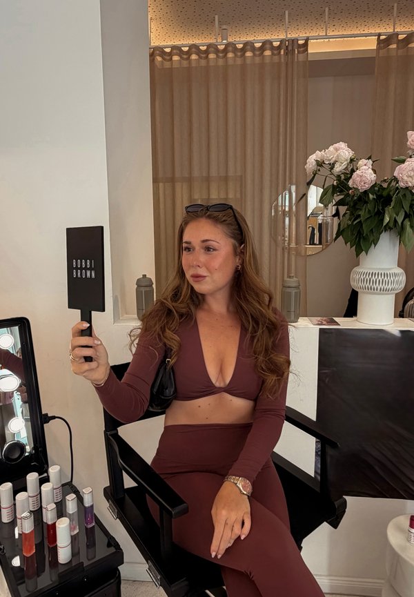 Frau in braunem Outfit sitzt auf einem Stuhl und hält einen Bobbi Brown Handspiegel in einem Make-up-Studio mit Lippenstiften und Blumen in der Nähe.